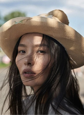 Close-up portrait of a woman wearing a straw hat with loose dark hair blowing across her face under natural light.