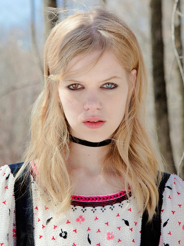 Close-up portrait of a young adult woman with blonde hair and unique heterochromia (different colored eyes) posing outdoors near bare trees.