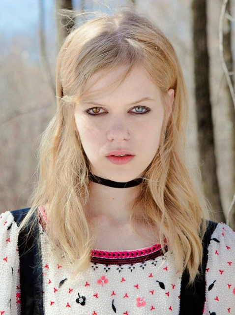 Close-up portrait of a young adult woman with blonde hair and unique heterochromia (different colored eyes) posing outdoors near bare trees.