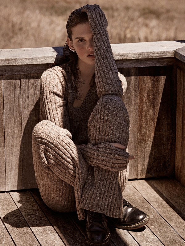 A young woman with dark hair sits cross-legged on a wooden deck, wearing a brown knit sweater and pants in an outdoor fashion shot.