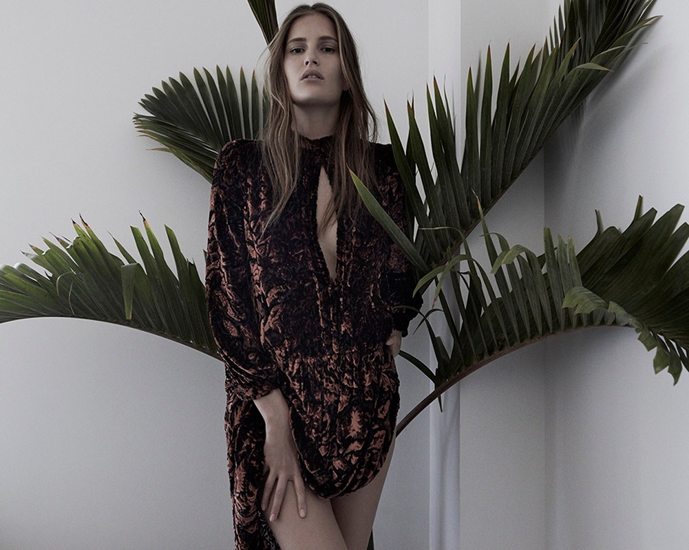 A young adult woman in a patterned dress stands indoors near a vibrant palm plant against a light wall, showcasing fashion and nature.