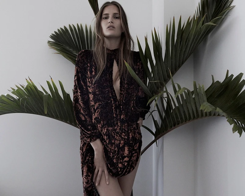 A young adult woman in a patterned dress stands indoors near a vibrant palm plant against a light wall, showcasing fashion and nature.