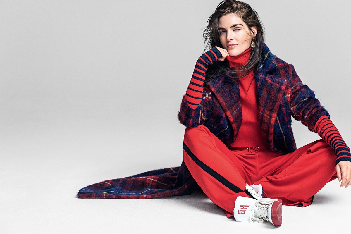 A female model sits in a studio, wearing a red sweater, pants with a navy stripe, a plaid coat, and white sneakers with red trim.