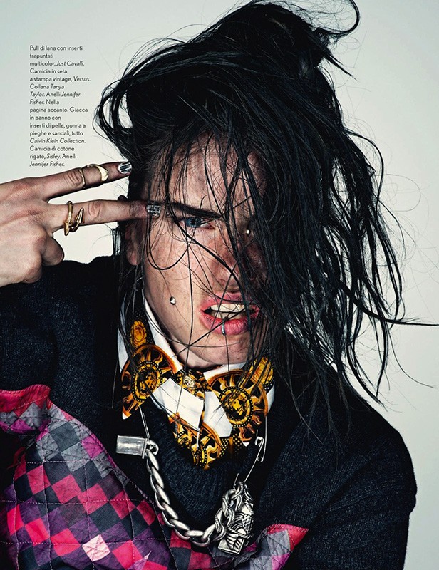 Person with dark hair and unique makeup in a fashion editorial, wearing a patterned bow tie and chain necklace, looking intensely at camera.