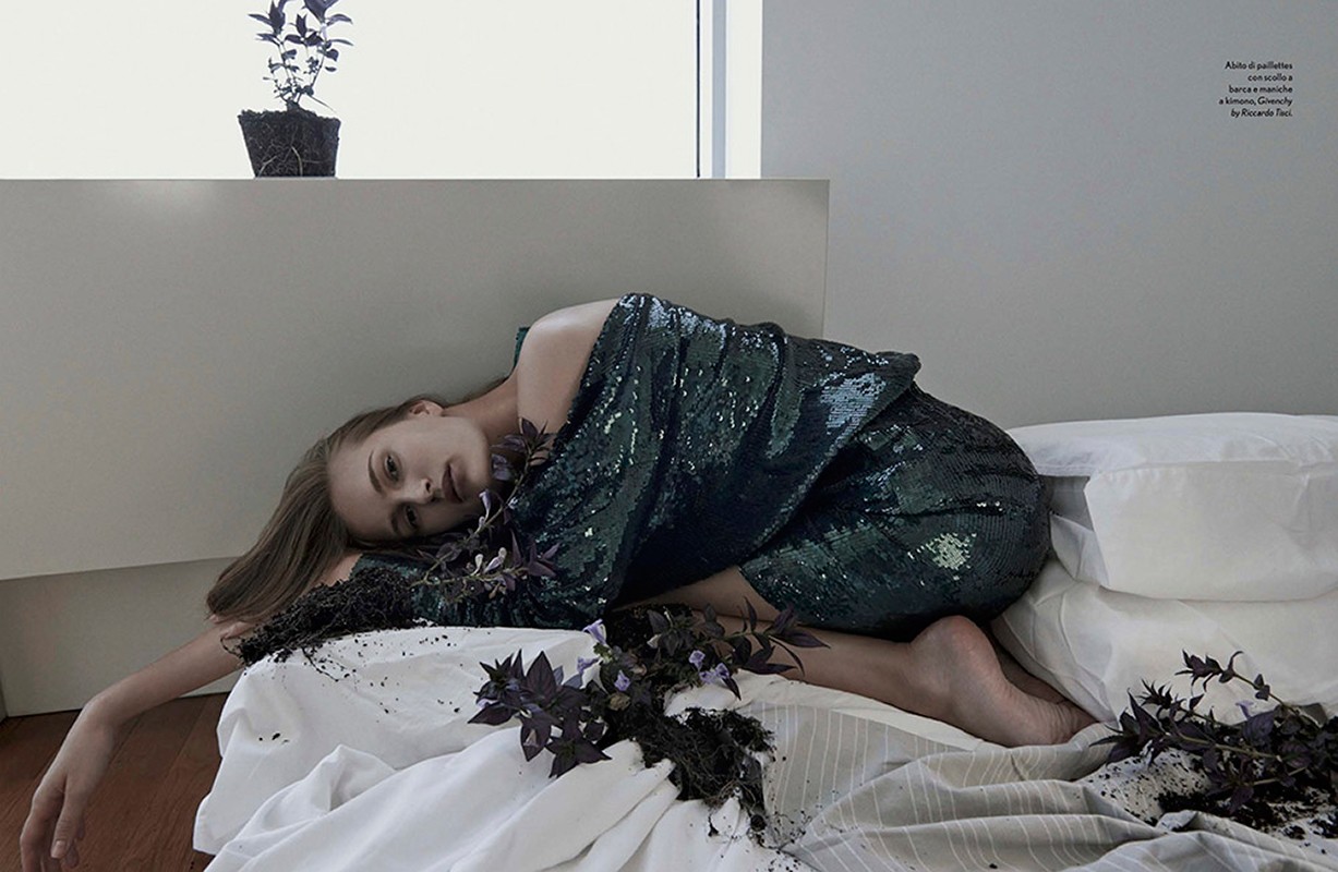 Young woman in a green sequin dress reclines on a white bed surrounded by purple herbs, with a potted plant in the soft-lit background.