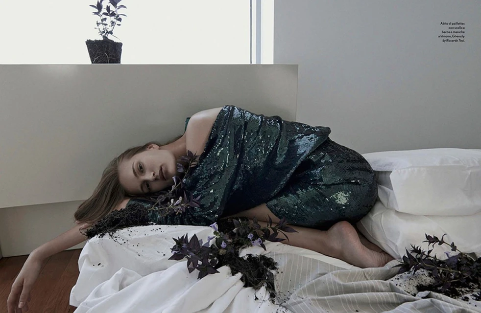 Young woman in a green sequin dress reclines on a white bed surrounded by purple herbs, with a potted plant in the soft-lit background.