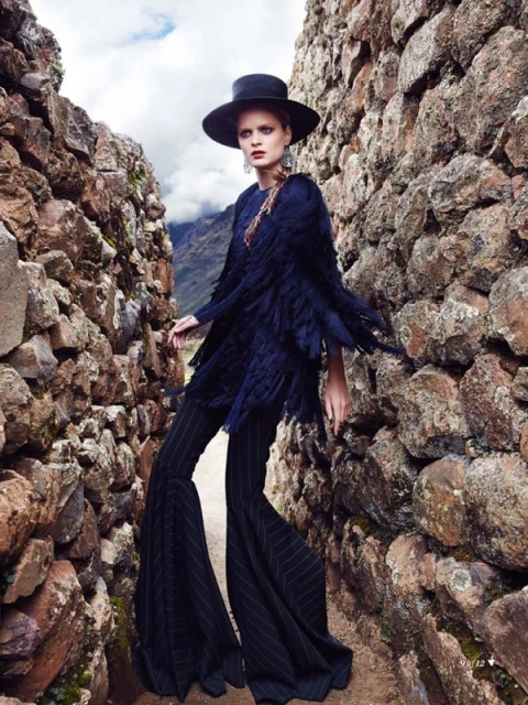 Fashion editorial shot of a woman in a dark blue fringed coat and striped pants walking through a stone alley with mountains in the distance.