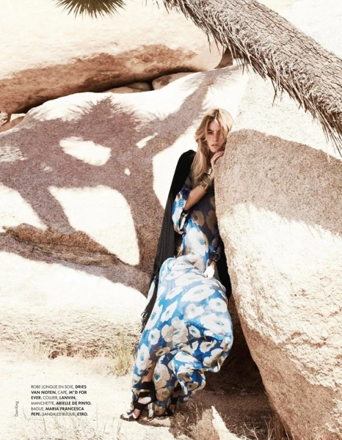 Woman in flowing floral dress poses amongst desert rocks, wearing a gold cuff bracelet, with a palm tree visible overhead.