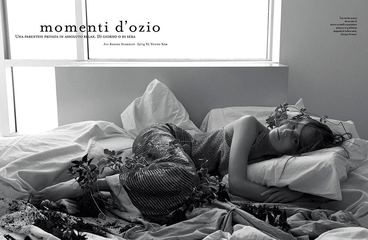 Woman in sparkling top and palazzo pants lies on white bedding in a dimly lit room decorated with plants near a bright window.