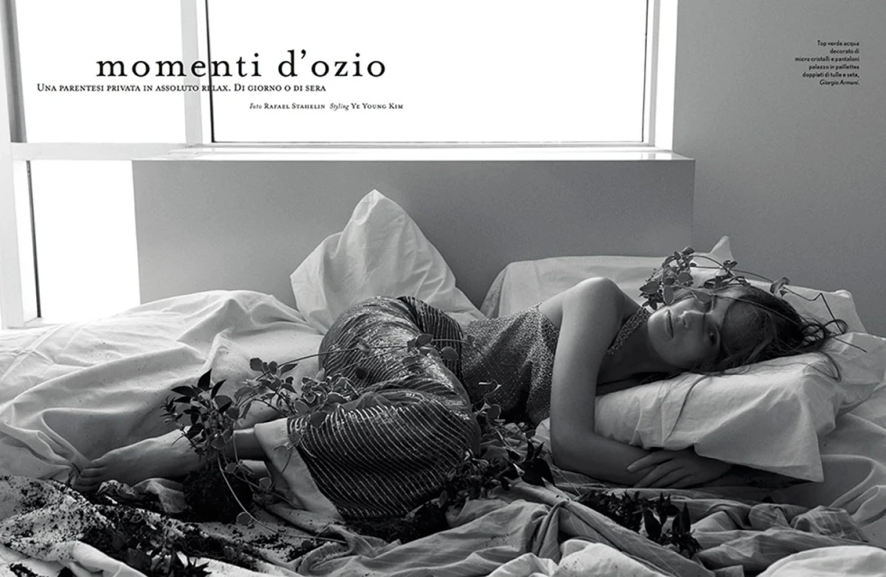 Woman in sparkling top and palazzo pants lies on white bedding in a dimly lit room decorated with plants near a bright window.