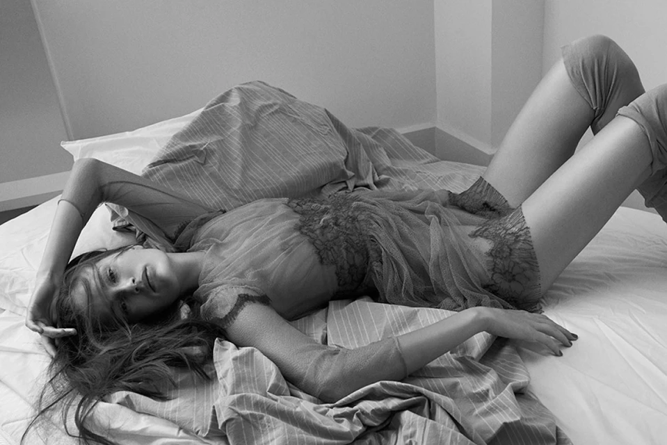 A woman with long brown hair lies on a bed with white sheets and striped blanket. She is wearing a sheer top and grey leggings.