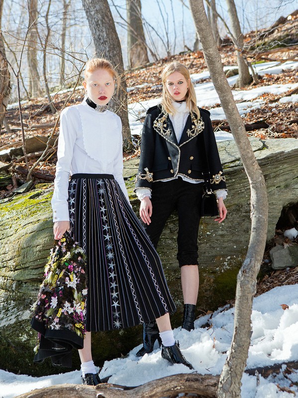 Two fair-skinned fashion models pose in a snowy forest setting, wearing contrasting black-and-white outfits, and looking at the camera.