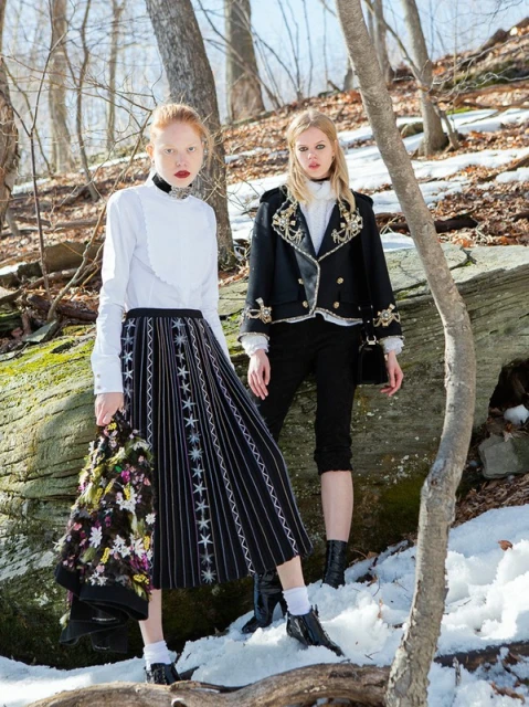 Two fair-skinned fashion models pose in a snowy forest setting, wearing contrasting black-and-white outfits, and looking at the camera.