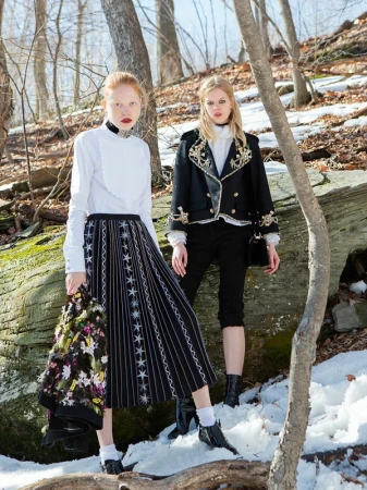 Two fair-skinned fashion models pose in a snowy forest setting, wearing contrasting black-and-white outfits, and looking at the camera.