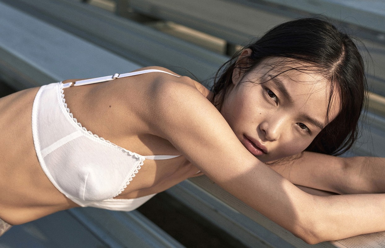 Close-up of a young Asian woman in a white bra lying on a wooden surface in soft, natural lighting, creating a fashion-forward editorial image.