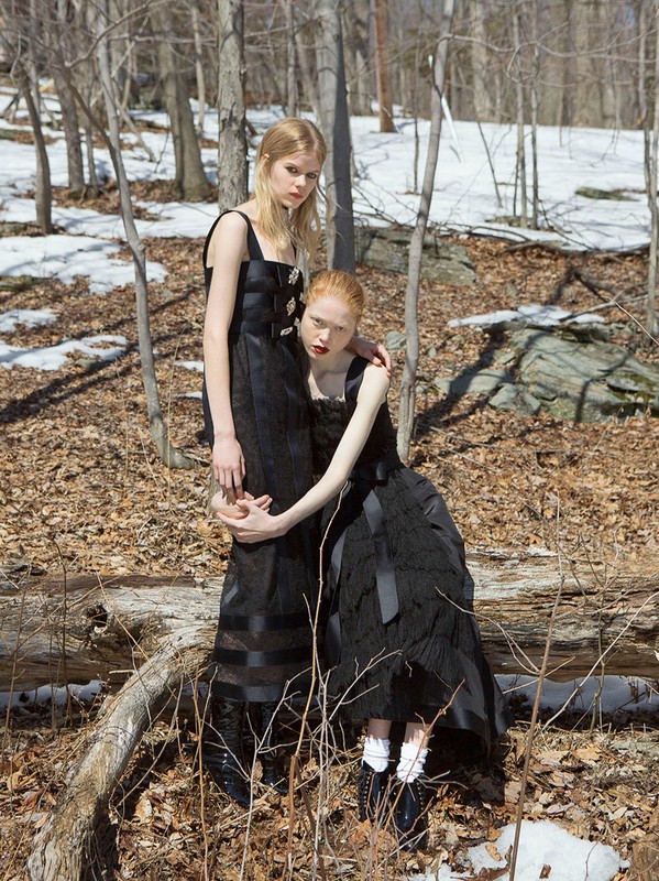 Two fair-skinned women in black dresses pose in a wooded area with fallen leaves and patches of snow, creating a striking fashion editorial.