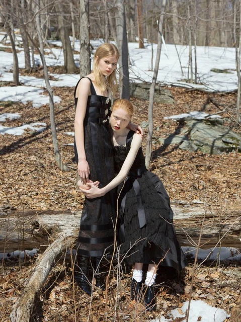 Two fair-skinned women in black dresses pose in a wooded area with fallen leaves and patches of snow, creating a striking fashion editorial.