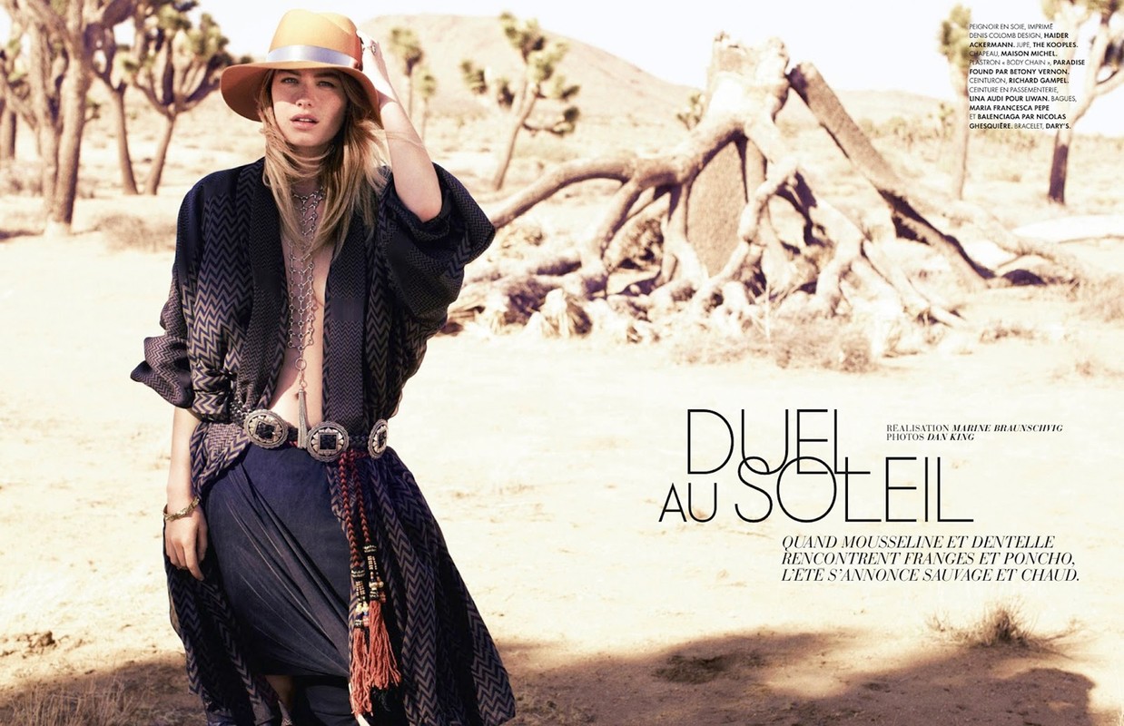 A woman wearing a hat and poncho poses in the desert against a backdrop of Joshua trees and arid landscape, captured in a fashion editorial style.