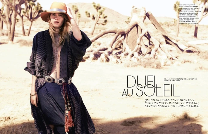 A woman wearing a hat and poncho poses in the desert against a backdrop of Joshua trees and arid landscape, captured in a fashion editorial style.