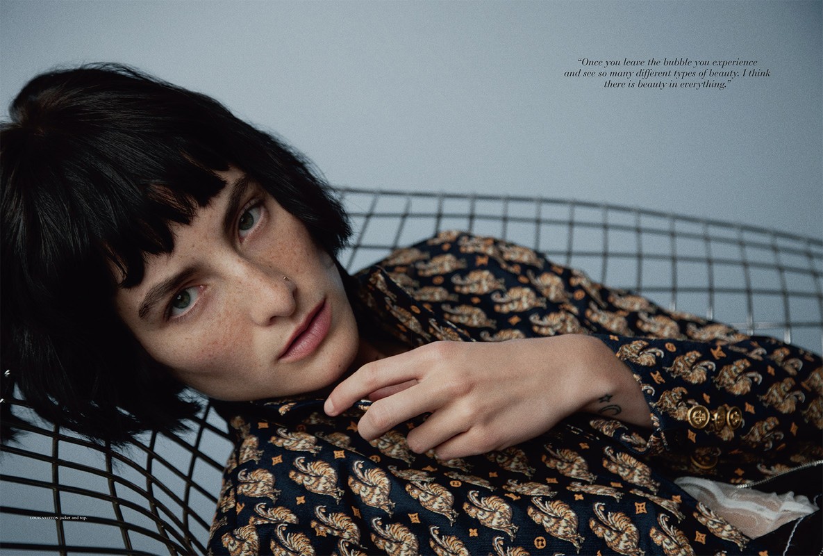 Portrait of a young woman with short dark hair wearing a patterned jacket, reclining in a metal chair indoors with a gray background.