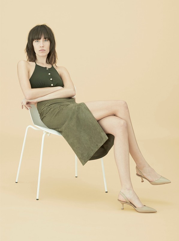 A woman in a green top and skirt sits on a chair in a studio with sling-back heels, arms crossed and legs crossed, in a full shot.