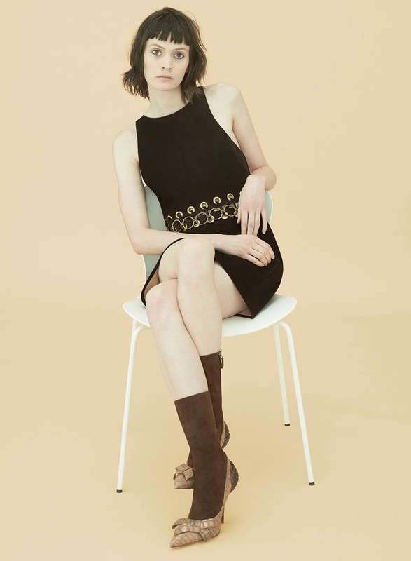 A woman with dark hair sits on a chair, wearing a black dress with a belt and brown boots in a studio setting.
