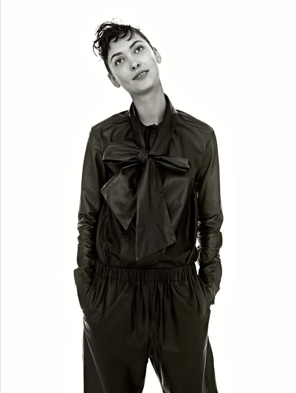 A woman poses indoors wearing a dark, shiny jumpsuit with a large bow. A medium shot captures her against a bright white background.
