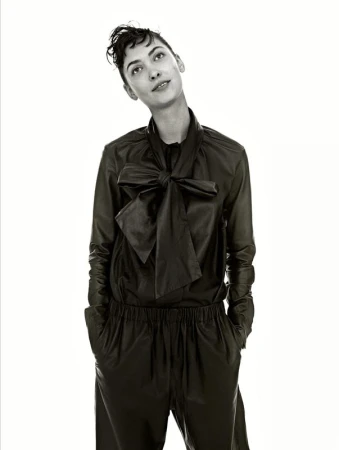 Portrait of a woman wearing a dark bow-collared shirt and trousers with hands in pockets against a plain background
