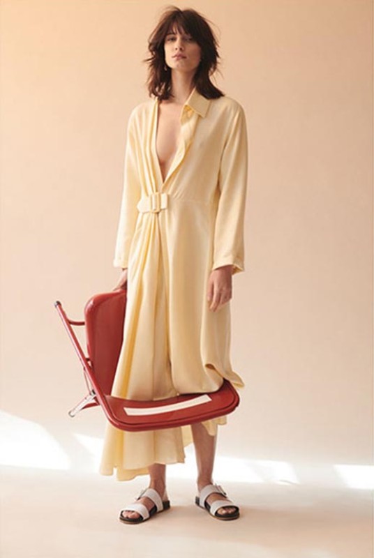 Fashion model wearing a yellow dress and white sandals stands in a studio holding a red chair in a fashion editorial photoshoot.