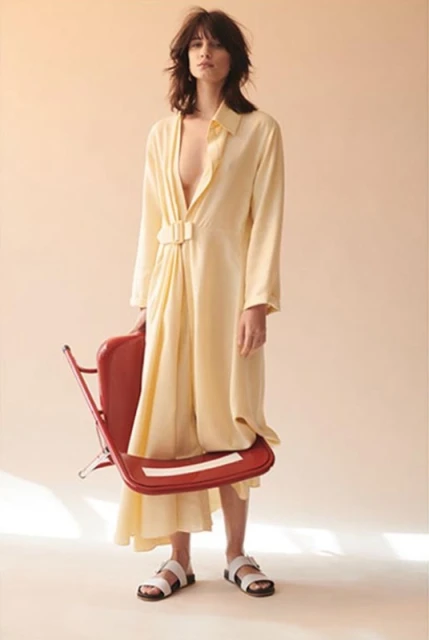 Fashion model wearing a yellow dress and white sandals stands in a studio holding a red chair in a fashion editorial photoshoot.