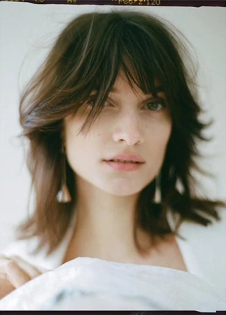 Close-up portrait of a woman with shoulder-length brown hair and bangs, wearing drop earrings, in a softly lit indoor setting.