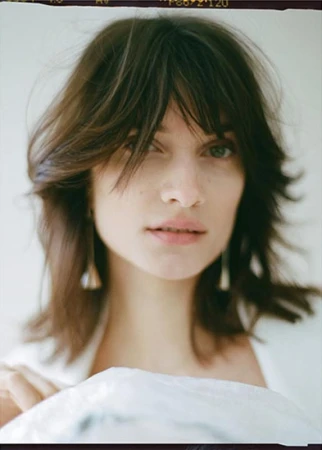 Close-up portrait of a woman with shoulder-length brown hair and bangs, wearing drop earrings, in a softly lit indoor setting.