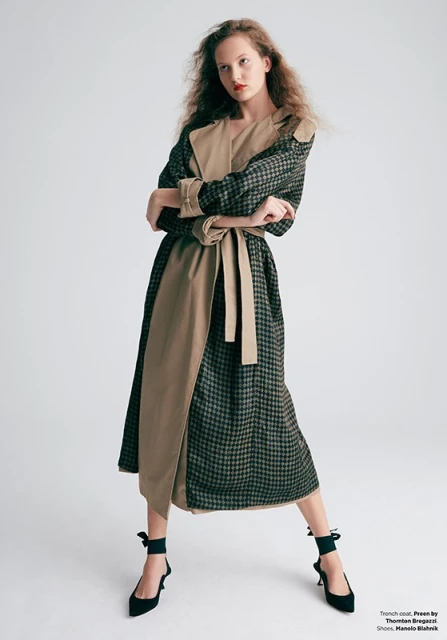 Fashion model in a modern trench coat and high heels poses in a photo studio against a plain background with soft, neutral lighting.