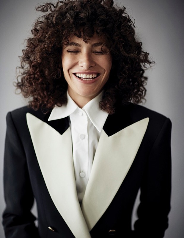 Smiling woman with curly hair wearing a black and white tuxedo blazer in a studio portrait.
