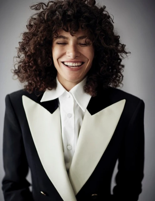 Smiling woman with curly hair wearing a black and white tuxedo blazer in a studio portrait.