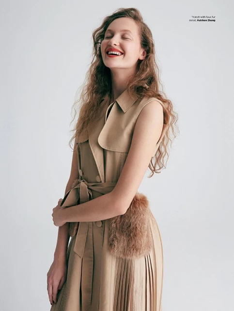 Smiling woman with closed eyes wearing a sleeveless tan dress with a fur accent against a plain white background in a studio setting.