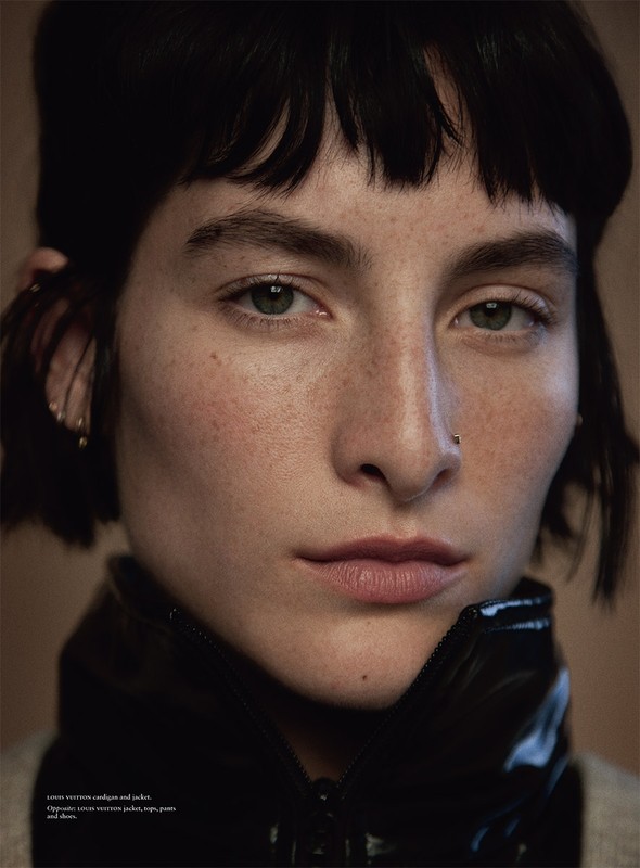 Close-up portrait of a woman with short dark hair, freckles, and green eyes, wearing a shiny black jacket against a neutral background.
