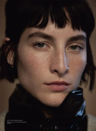 Close-up portrait of a model with dark short hair, freckles, and a black shiny zipped collar against a neutral background.