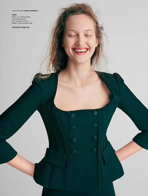 Smiling woman in a dark green jacket and skirt, hands on hips, with fair skin and wavy hair against a light background in a studio.