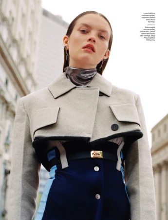 Model wearing a cropped grey jacket and blue dress with a metallic neckpiece outdoors with tall buildings.