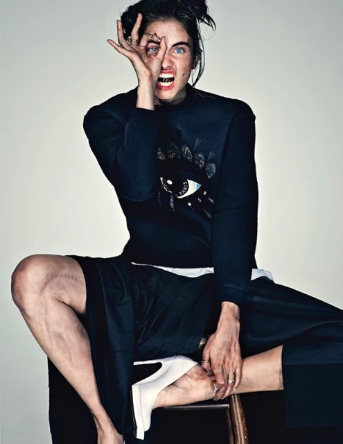 A striking portrait of a young adult female model sitting on a stool, striking a pose with her hand over one eye in a fashion editorial.