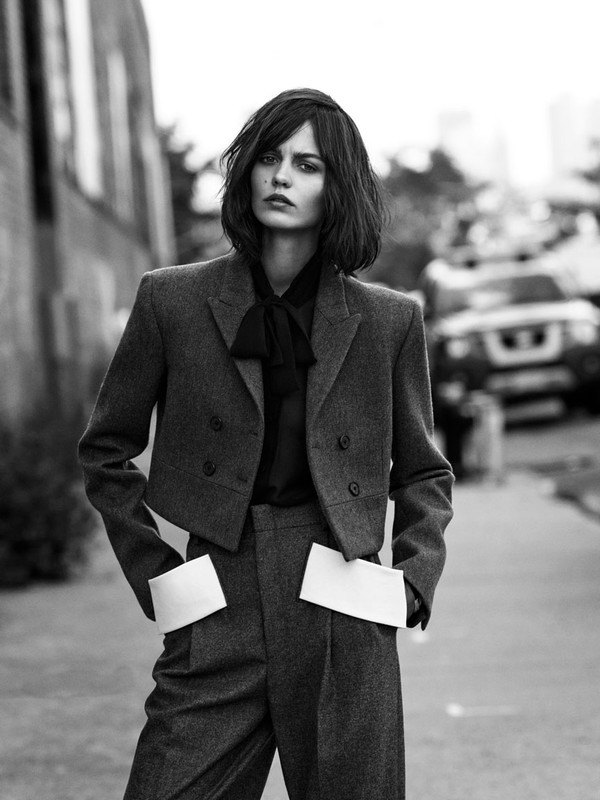 Black and white fashion shot of an adult woman in a dark blazer and trousers, with a serious expression, in an urban setting.