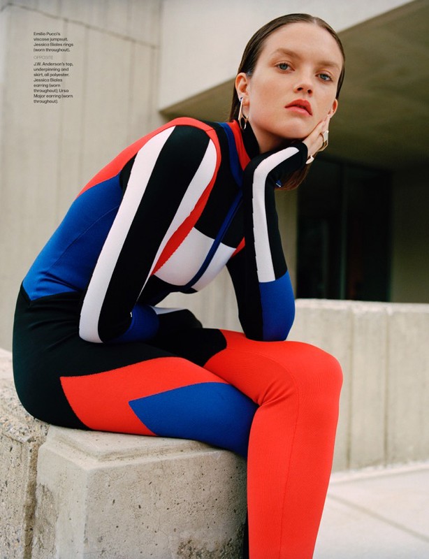 A fashion model in a color-blocked bodysuit sits outdoors against a concrete wall, with muted natural lighting and a minimalist aesthetic.
