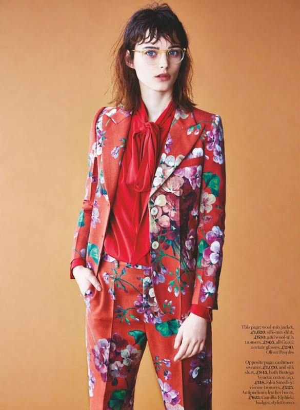 Fashion editorial shot: Brunette woman with glasses poses in a floral patterned suit and red blouse against an orange background.