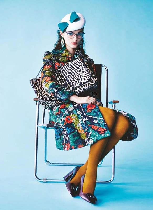 A stylish woman in a patterned coat sits in a chair, holding a matching handbag, against a light blue backdrop in a fashion setting.