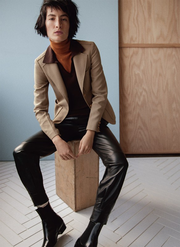 Brunette woman in a beige blazer, brown turtleneck, black leather pants and boots sits on a beige cube against a neutral backdrop.