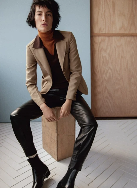 Brunette woman in a beige blazer, brown turtleneck, black leather pants and boots sits on a beige cube against a neutral backdrop.