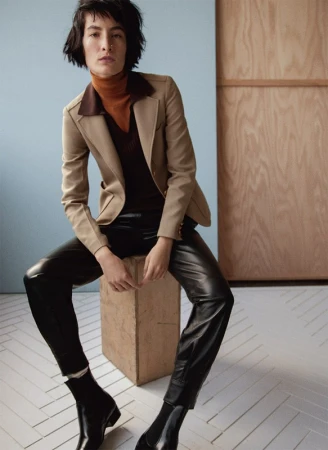 Brunette woman in a beige blazer, brown turtleneck, black leather pants and boots sits on a beige cube against a neutral backdrop.