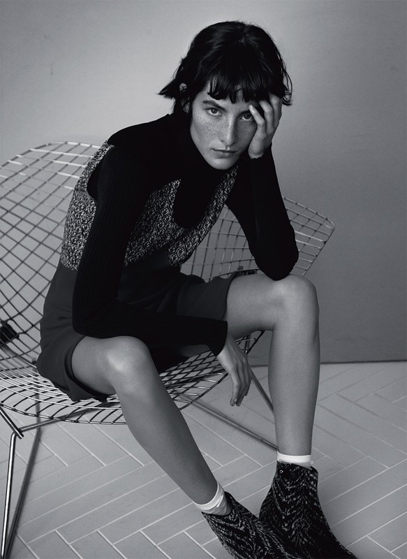 Black and white portrait of a woman with short dark hair sitting on a metal chair, gazing at the camera in a photo studio.