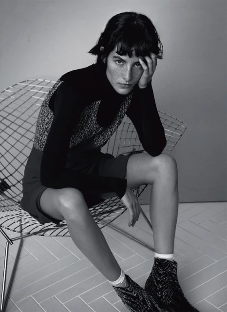 Black and white portrait of a woman with short dark hair sitting on a metal chair, gazing at the camera in a photo studio.